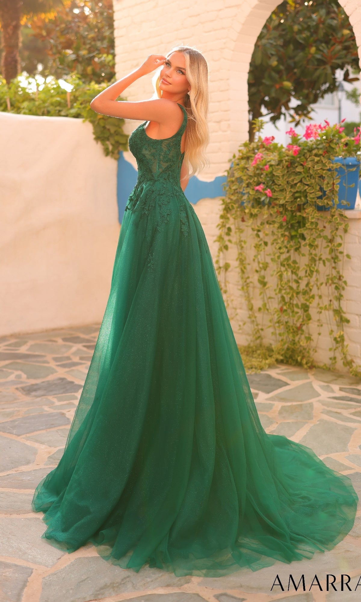 A woman wearing the Amarra 88201 long prom dress, featuring a sleeveless green design and embroidered corset bodice, stands outdoors on a stone path with greenery and white walls in the background, looking over her shoulder.